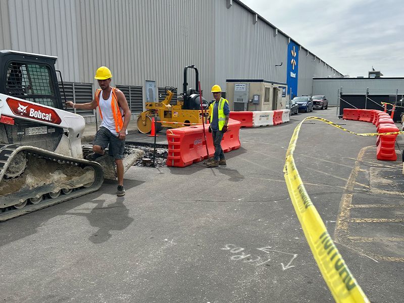 Two construction workers by a Bobcat, repairing asphalt, with caution barriers.