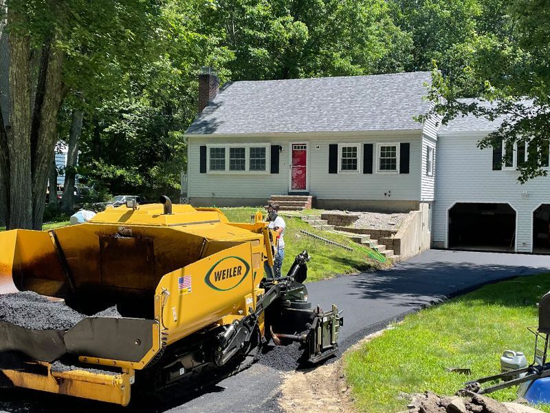 Yellow paving machine at a house driveway with a red door and open garage on a sunny day