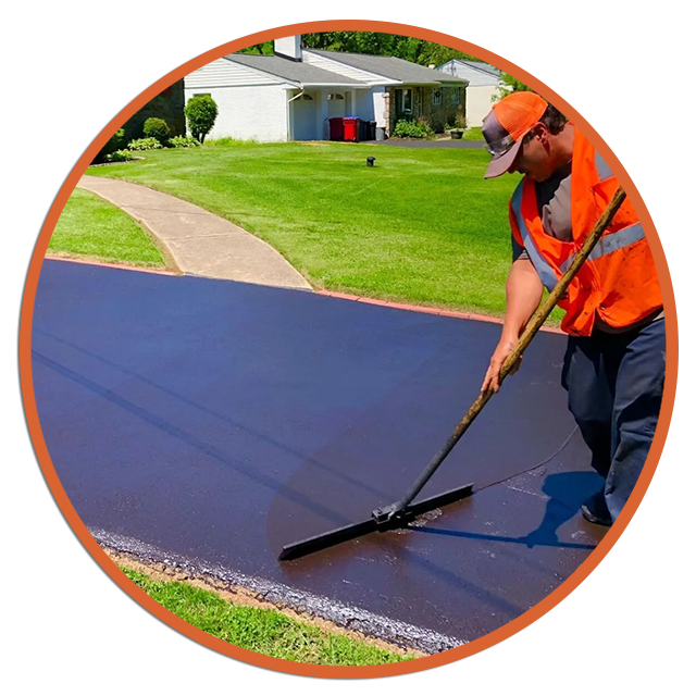 Worker spreading black sealant on a driveway with a squeegee, wearing an orange safety vest and hat
