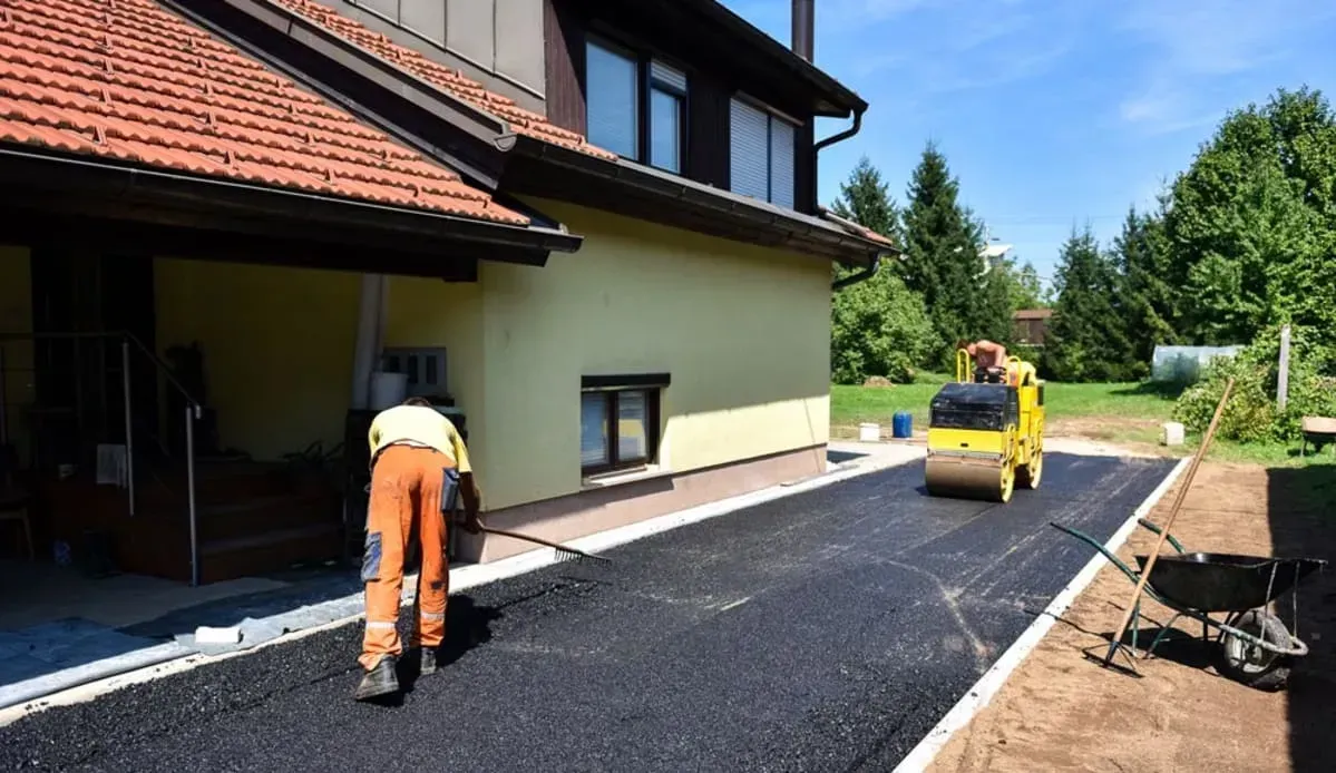 Workers paving a driveway in front of a yellow house with a red tiled roof. A roller machine compacts asphalt.