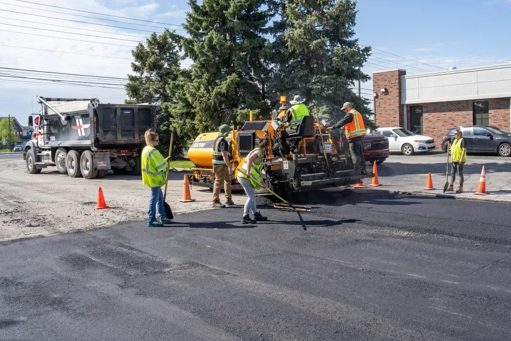 Road construction crew paving asphalt road with machinery, dump truck, and safety cones.