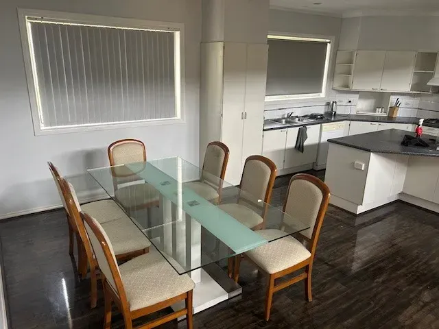 Dining area with a large glass table and eight upholstered chairs, adjacent to a kitchen with white cabinets and dark countertops.
