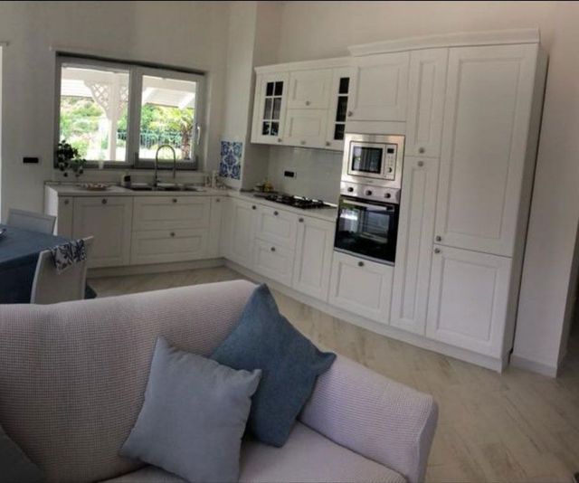 Cucina moderna bianca con elettrodomestici a incasso, finestra sopra il lavello e divano grigio in primo piano.