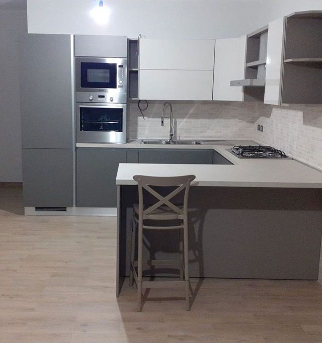 Cucina moderna grigia con isola, forno a incasso, lavello e una sedia singola di fronte.