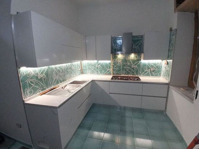 Cucina moderna a L di colore bianco con paraschizzi in piastrelle verdi, illuminazione sottopensile e pavimento piastrellato.
