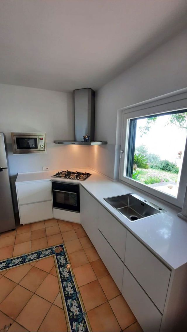 Cucina moderna bianca con pavimento in piastrelle di terracotta, piano cottura, lavello e ampia finestra con vista sul verde.