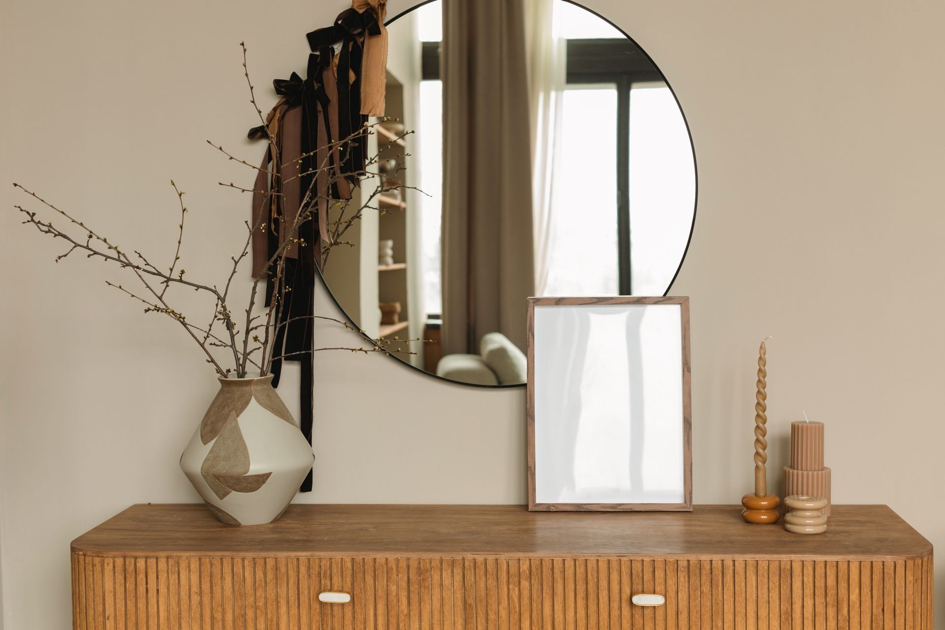 In una stanza minimalista, troviamo una cassettiera in legno con specchio rotondo, un vaso di rami, una stampa incorniciata e delle candele.