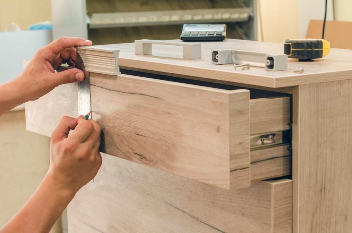 Mani che misurano con un metro a nastro il frontale di un cassetto in legno chiaro di un mobile da cucina.