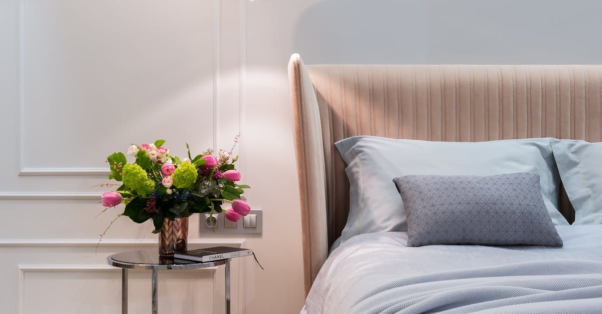 Camera da letto con testiera imbottita beige, lenzuola blu e un piccolo comodino con un colorato mazzo di fiori.