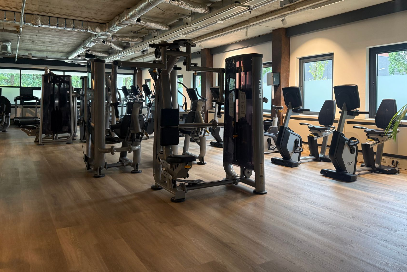 Lege fitnessruimte met fitnessapparaten en grote ramen op een houten vloer.