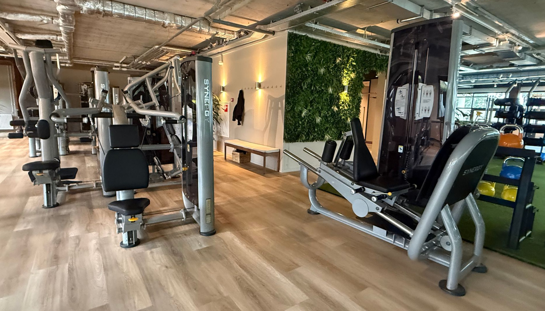 Modern fitnesscentrum met gewichtsapparaten, banken, houten vloeren en een groene plantenwand.