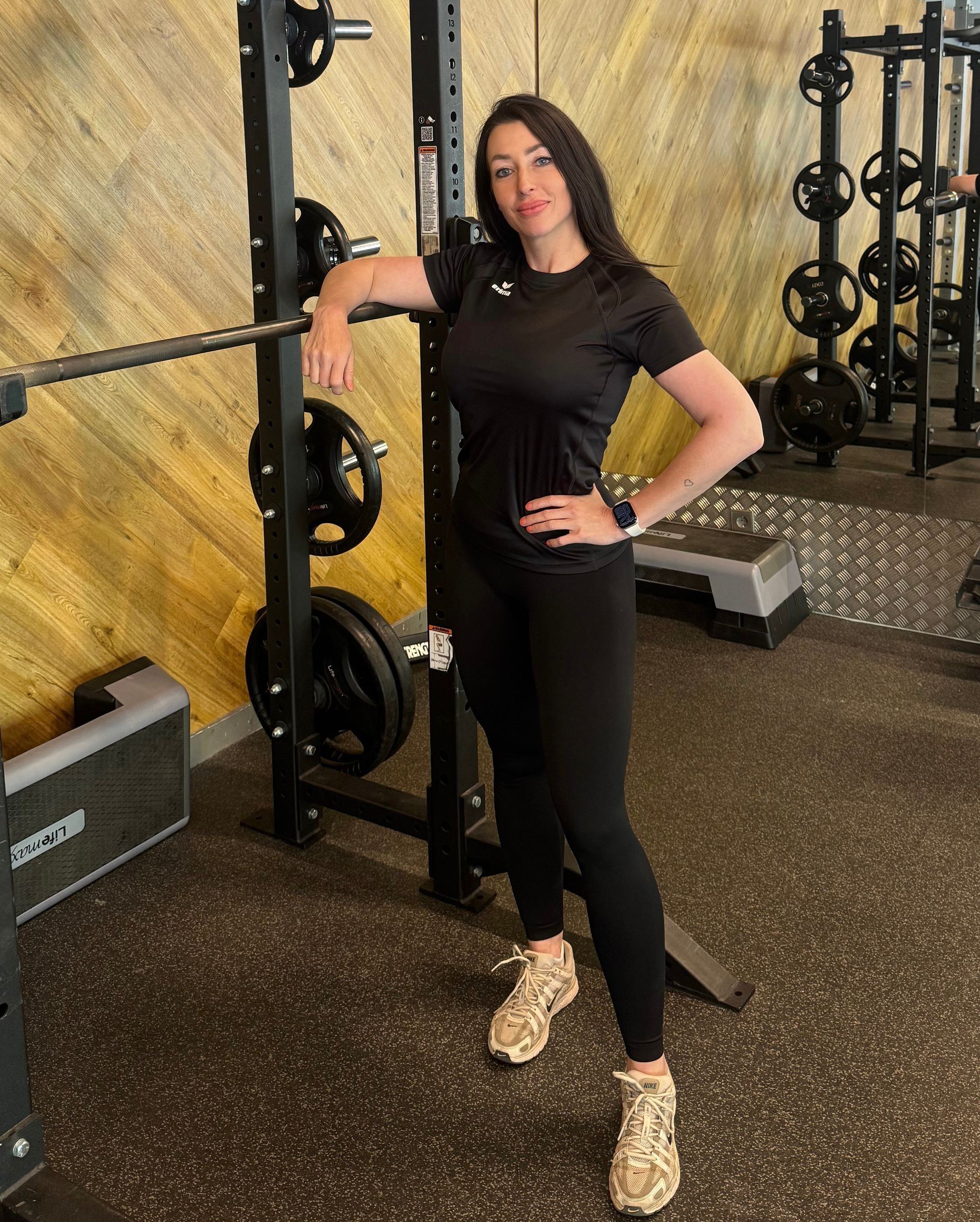 Vrouw poseert in een sportschool, leunend tegen een squat rack met één hand in haar zij.