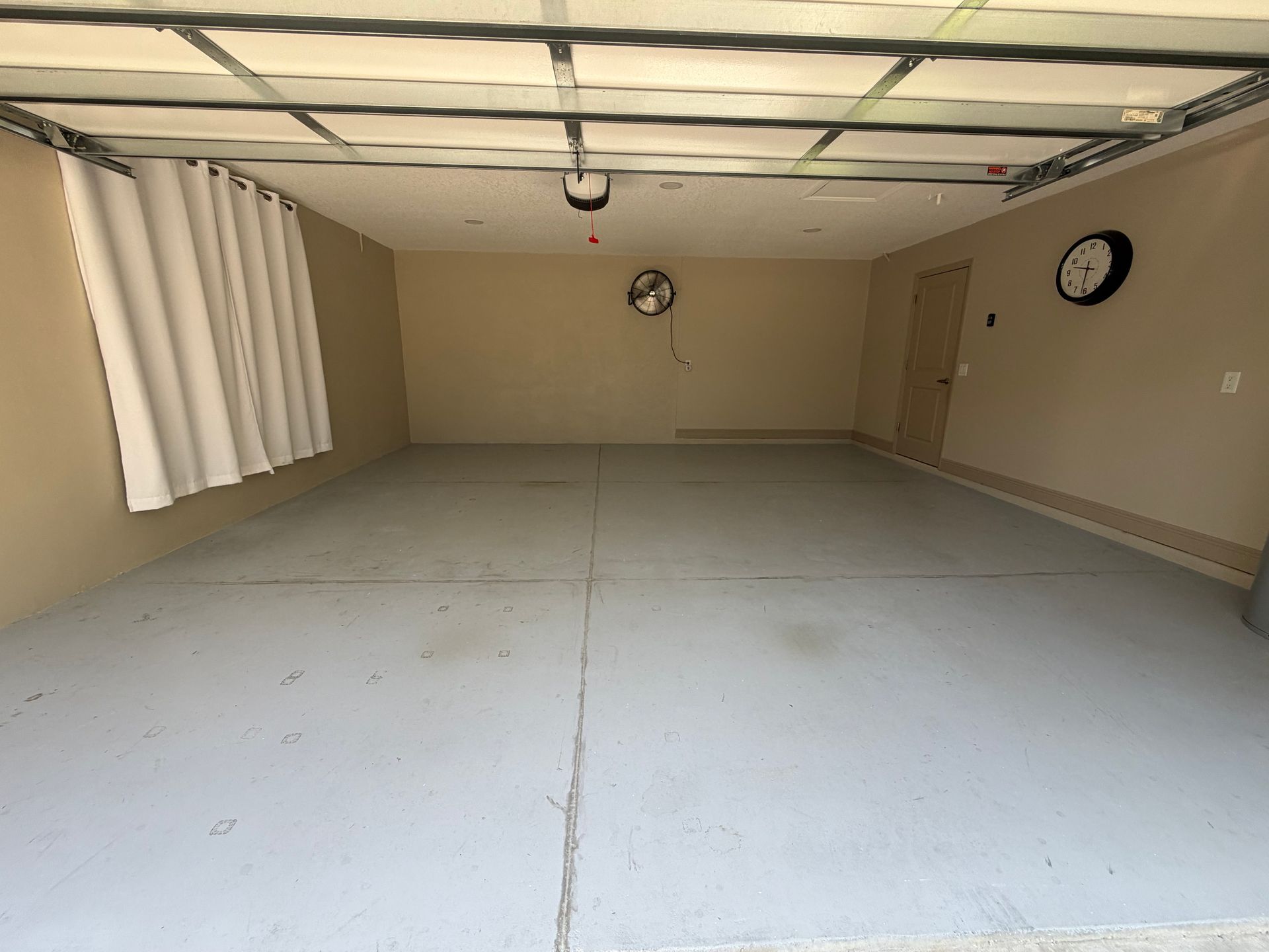 Empty garage with white floor, beige walls, curtained window, and two wall clocks.