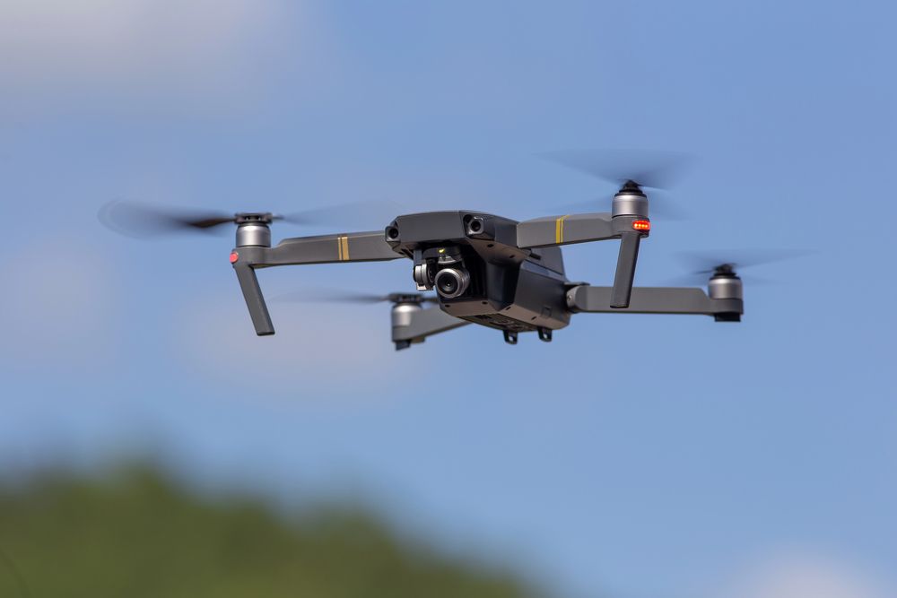 Dron en el aire, cuerpo gris oscuro, cuatro hélices girando, cámara negra, contra el cielo azul.
