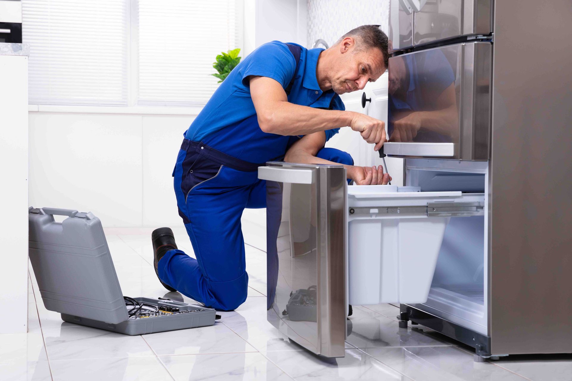 Technician in blue overalls repairing a stainless steel refrigerator in a kitchen
