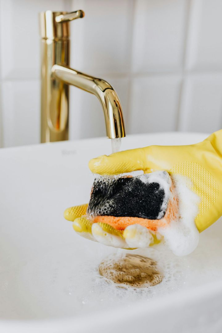Gloved hand scrubbing a dirty sponge under a running kitchen faucet