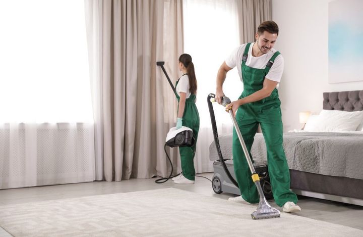 Two cleaners in green overalls vacuuming a bright bedroom with a bed and curtains