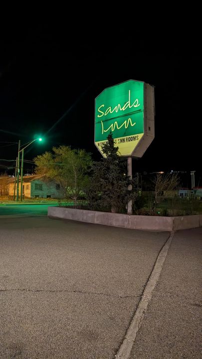 Nighttime roadside sign for Sandra Lodge glowing green above a low wall and trees.