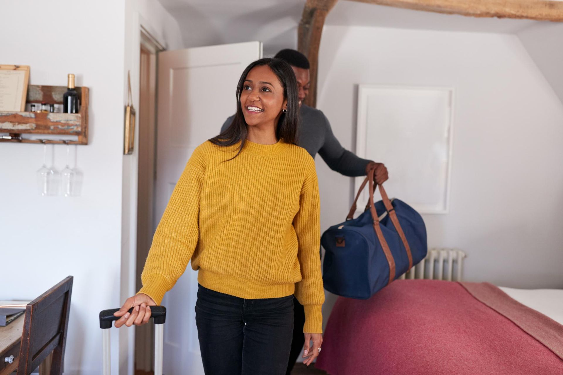 Woman in yellow sweater smiling while carrying a duffel bag in a bedroom doorway