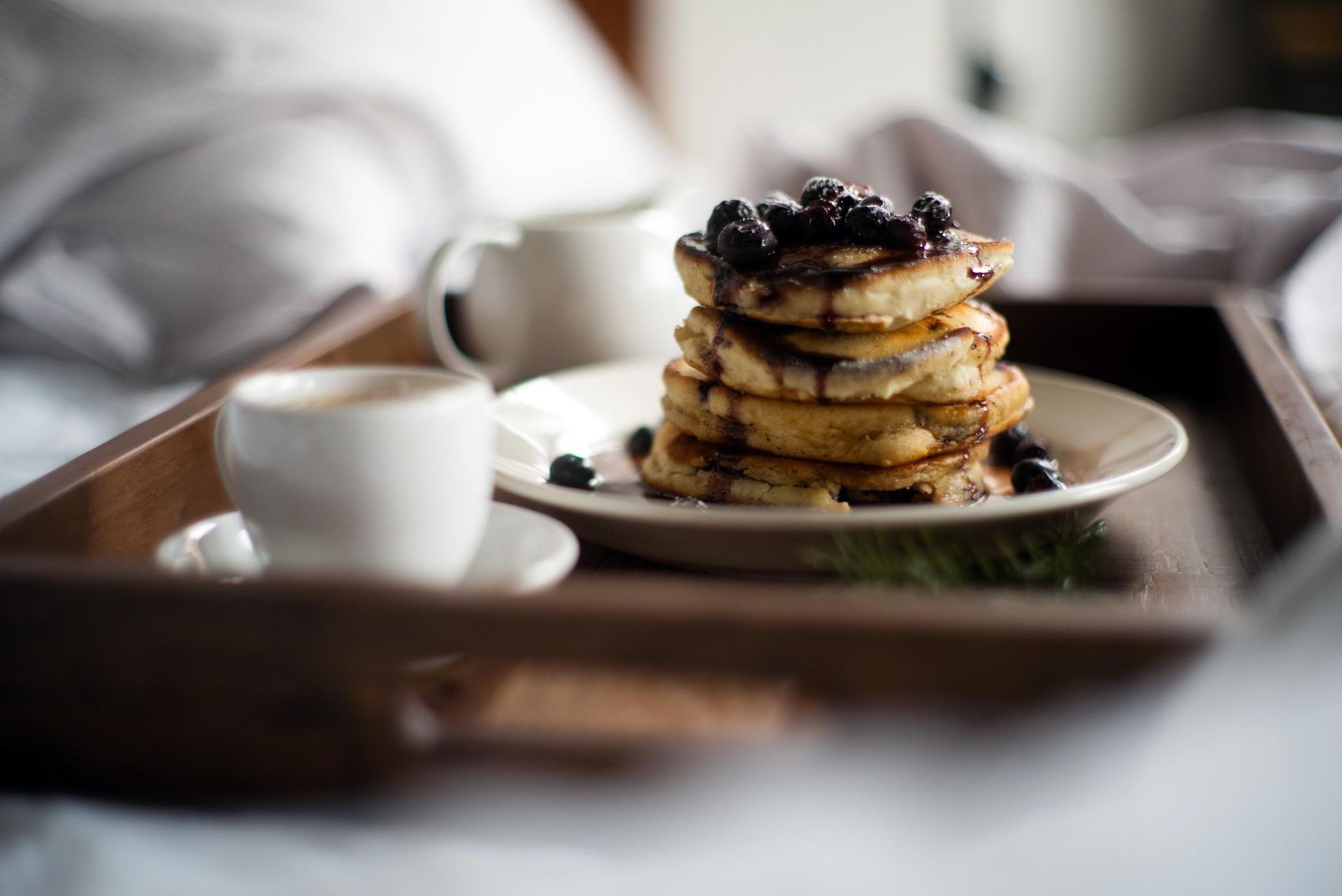 Stack of pancakes with berries on a tray beside a white coffee cup on a bed tray