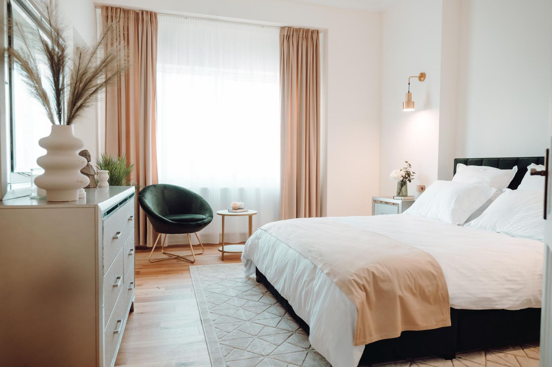Bright bedroom with a white bed, beige curtains, black chair, and dresser by a window