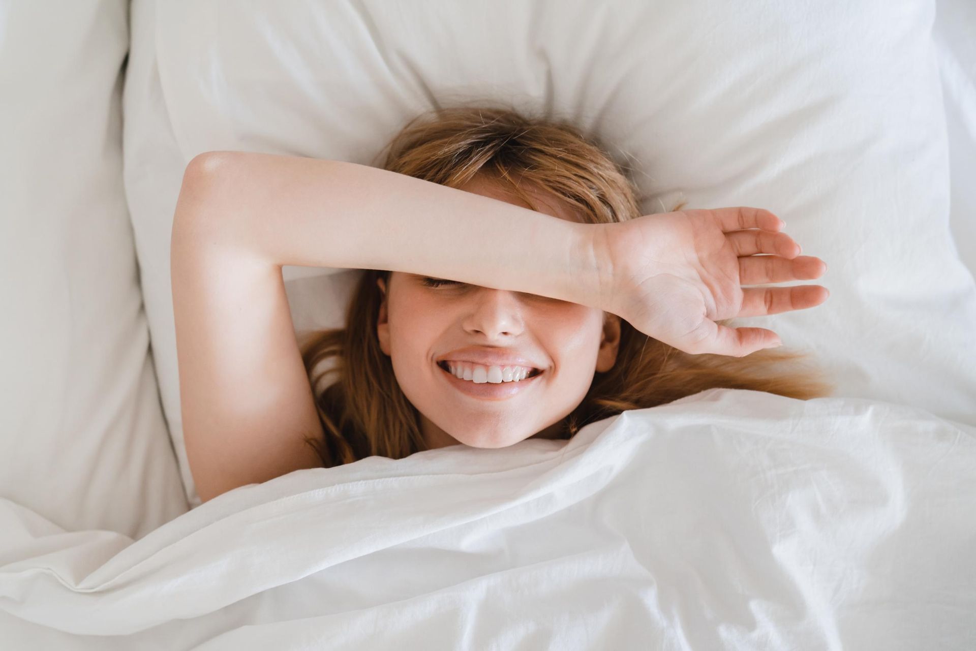 Smiling person lying in bed under white sheets, shielding eyes from bright light.