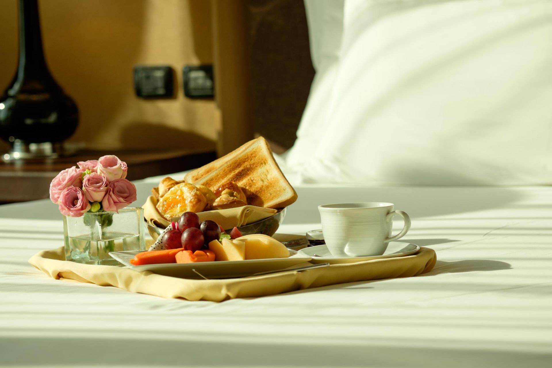 Breakfast tray with toast, fruit, coffee, and pink flowers on a bed