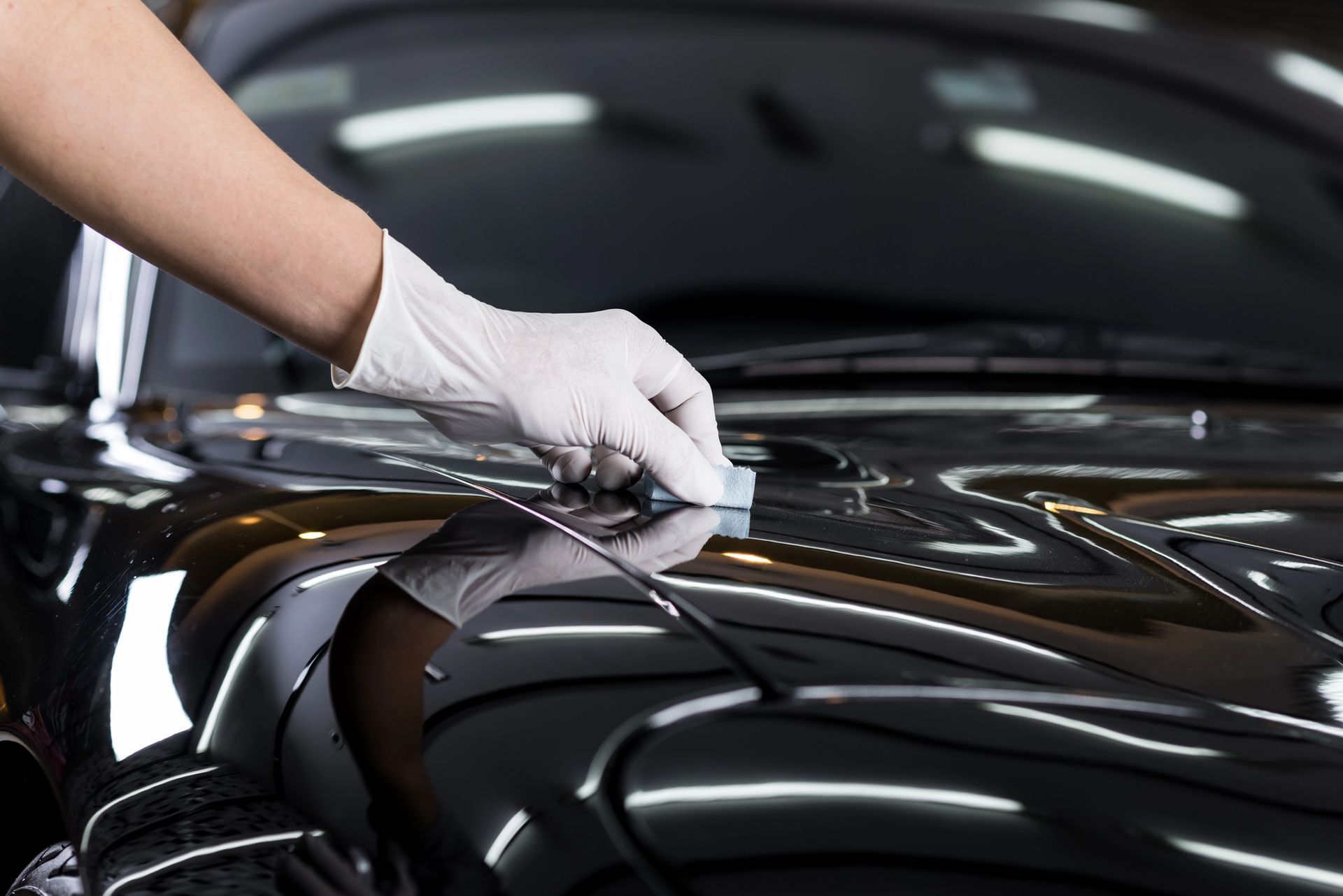 Mano guantata che applica uno strumento al cofano di un'auto nera lucida durante la fase di detailing.