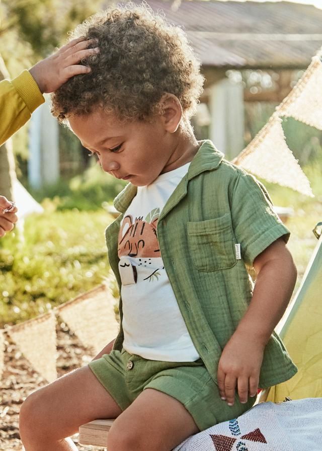 Ragazzo con maglietta verde e pantaloncini, con una maglietta bianca, all'esterno, mano sulla testa.