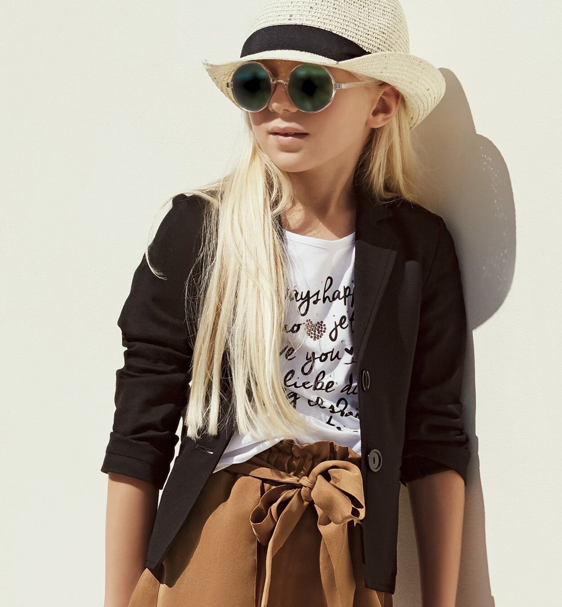 Ragazza con occhiali da sole, blazer, cappello e pantaloncini marroni, appoggiata a un muro bianco.