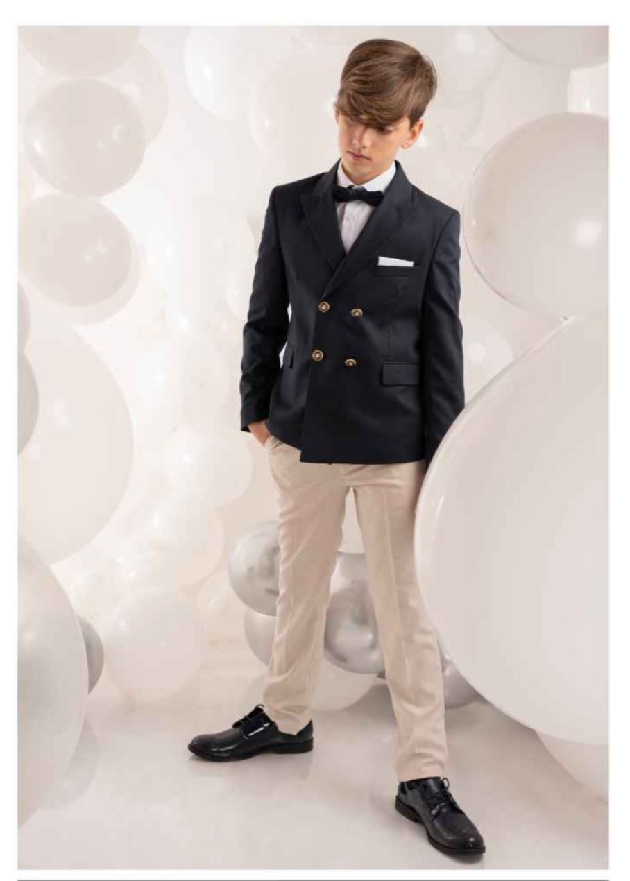 Un ragazzo con giacca blu navy e pantaloni color cuoio posa contro palloncini bianchi.