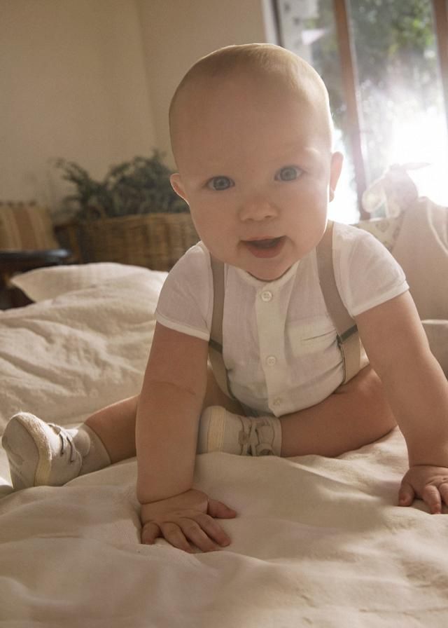 Un bambino è sdraiato sul letto, indossa una tutina bianca e delle bretelle e guarda la telecamera.
