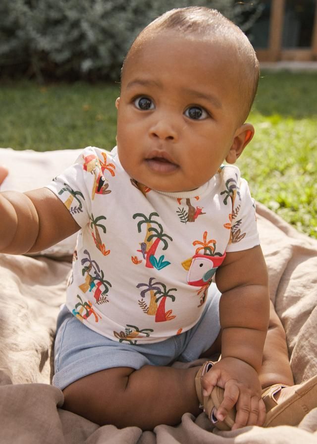 Un bambino indossa una maglietta bianca con disegni colorati e pantaloncini blu ed è seduto su una coperta all'aperto.