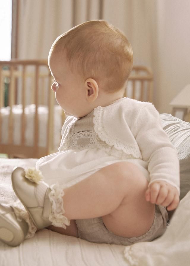 Neonato con vestito bianco e cardigan, seduto su un letto, che guarda di lato.