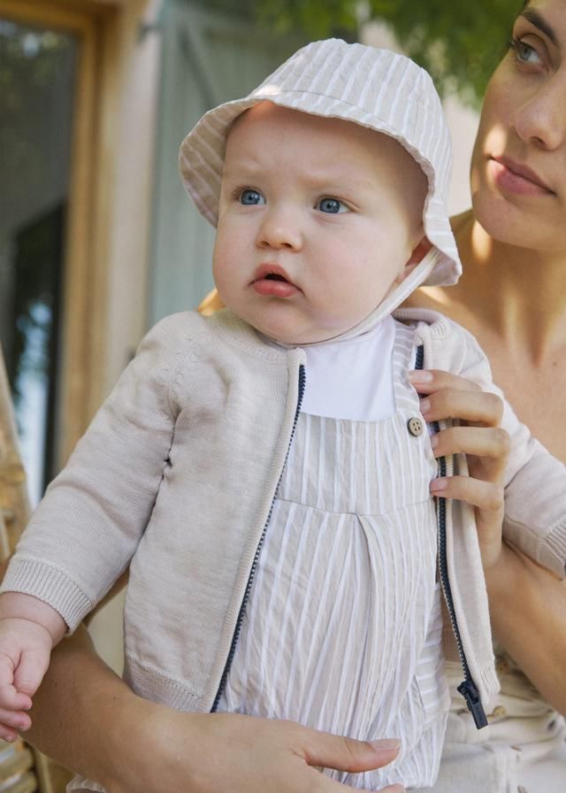 Un bambino con un vestito a righe e un cappellino da sole, tenuto in braccio da una persona dalla pelle chiara, in un ambiente esterno.