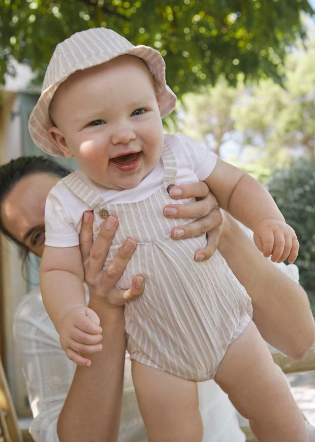 Un bambino sorridente, con una tuta a righe e un cappello da sole, viene tenuto in braccio da una persona all'esterno.