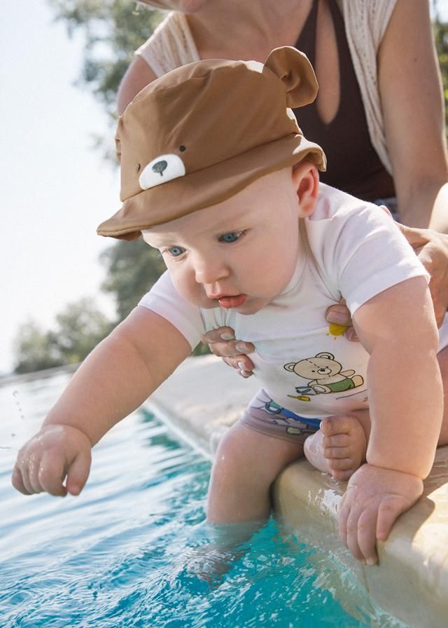 Un bambino che indossa un cappello a forma di orso tocca l'acqua di una piscina, tenuto in braccio da un adulto.