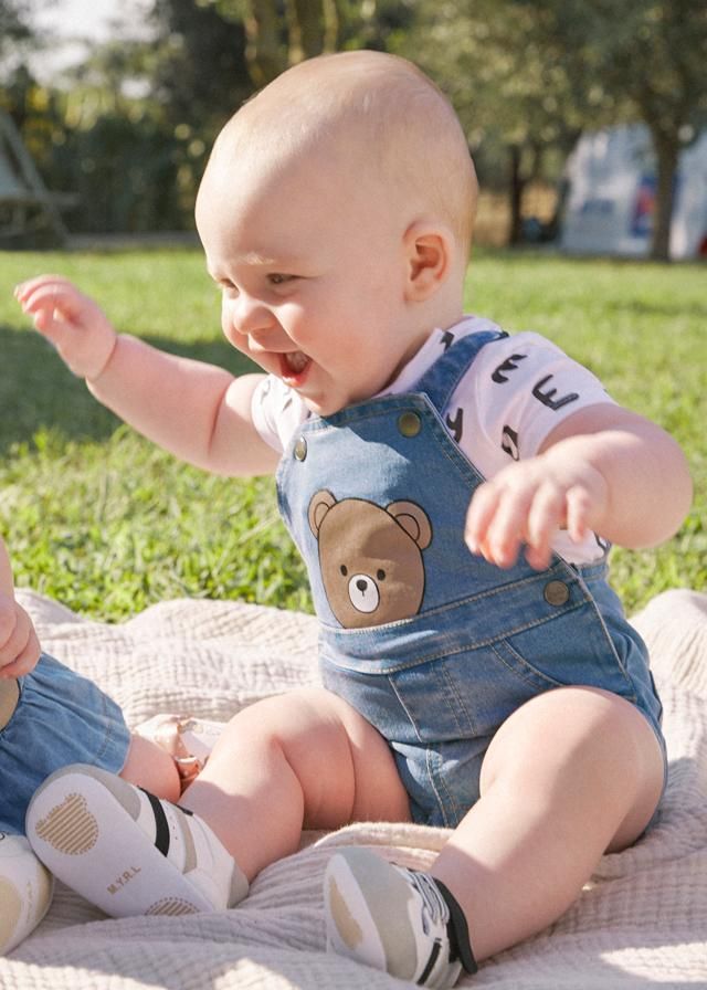 Un bambino con una salopette di jeans e la stampa di un orso è seduto su una coperta all'aperto, sorridente e con le braccia alzate.