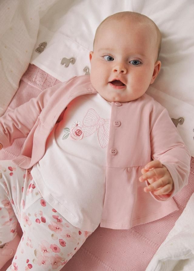 Neonato con abitino rosa, sorridente, sdraiato su una coperta bianca e coperta rosa.