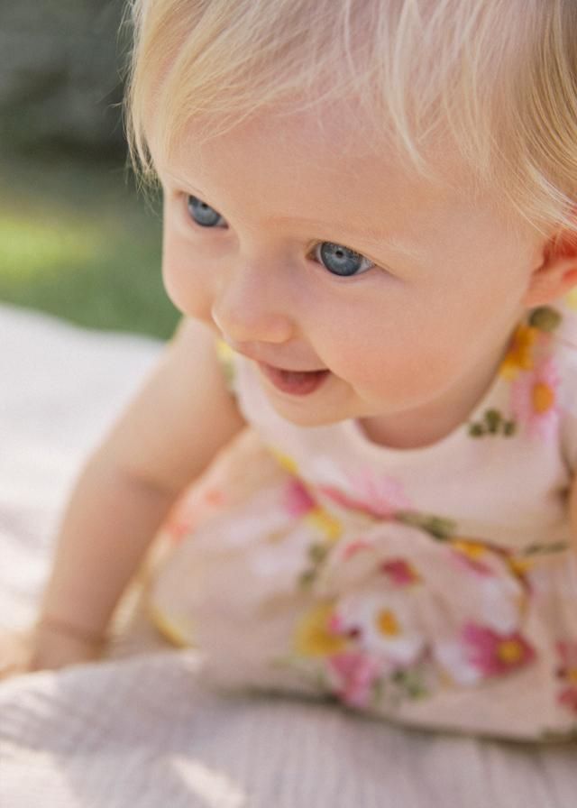 Una bambina bionda con gli occhi azzurri sorride, indossa un vestito a fiori ed è seduta su una coperta all'aperto.