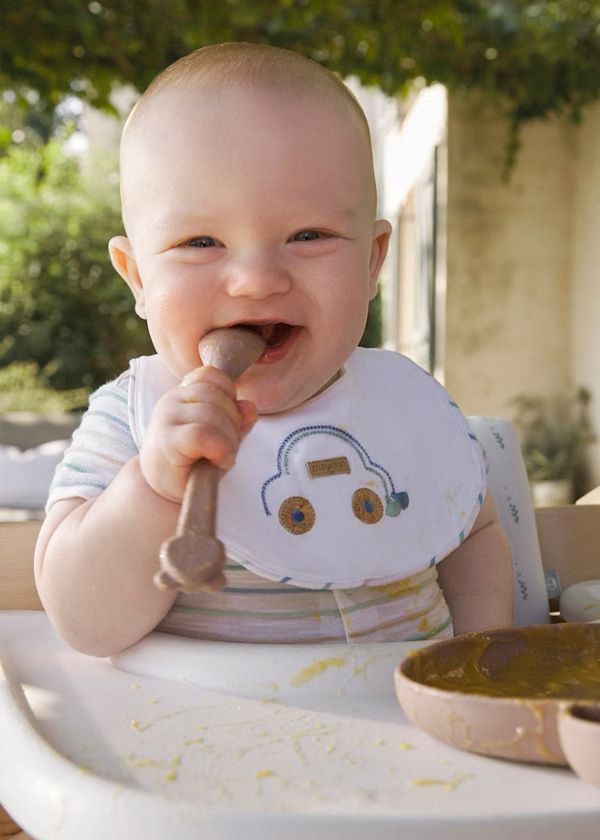 Un bambino nel seggiolone, sorridente, che mastica un cucchiaio di legno e indossa un bavaglino con un'auto.