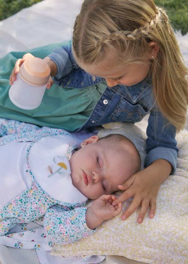 Una bambina con i capelli intrecciati osserva il neonato addormentato, che tiene in mano il biberon ed è appoggiato su una coperta.