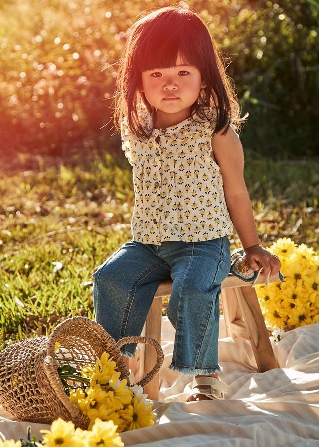 Una ragazza è seduta su uno sgabello di legno all'aperto, con indosso un top a fiori e jeans. È illuminata dal sole, con dei fiori e un cesto vicino.