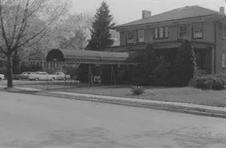 Black-and-white street view of a two-story house with a covered driveway and bare trees.
