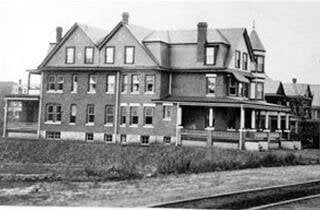 Black-and-white photo of a large brick building with a porch beside railroad tracks and an open field