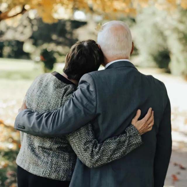 Two people standing with arms around each other, walking outdoors in an autumn setting with yellow leaves.