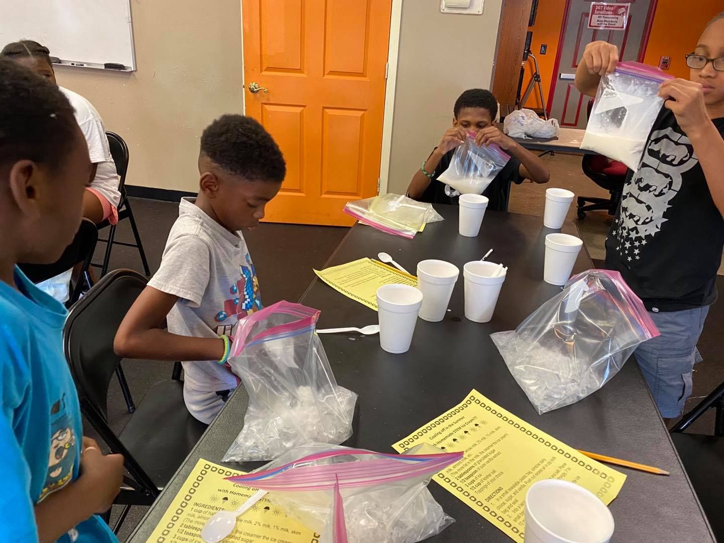 Students in a classroom perform a science experiment using plastic bags, ice, and white powder at a table with instructions.