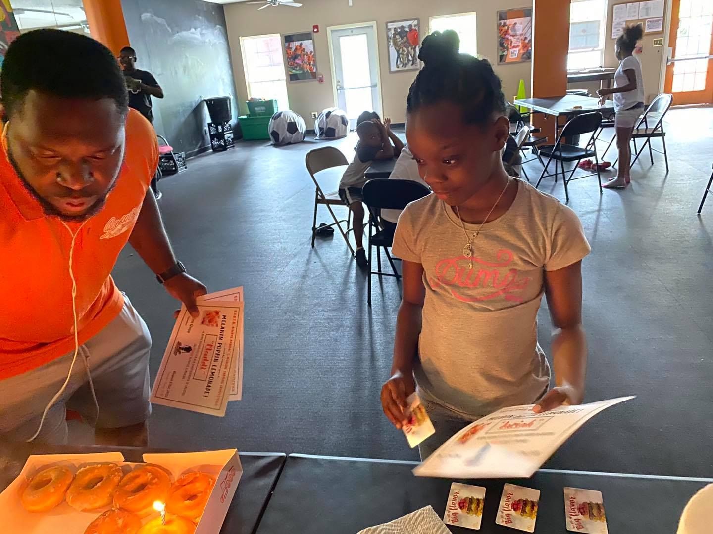 An instructor and a child stand at a table, holding awards beside a box of donuts in a multipurpose room.