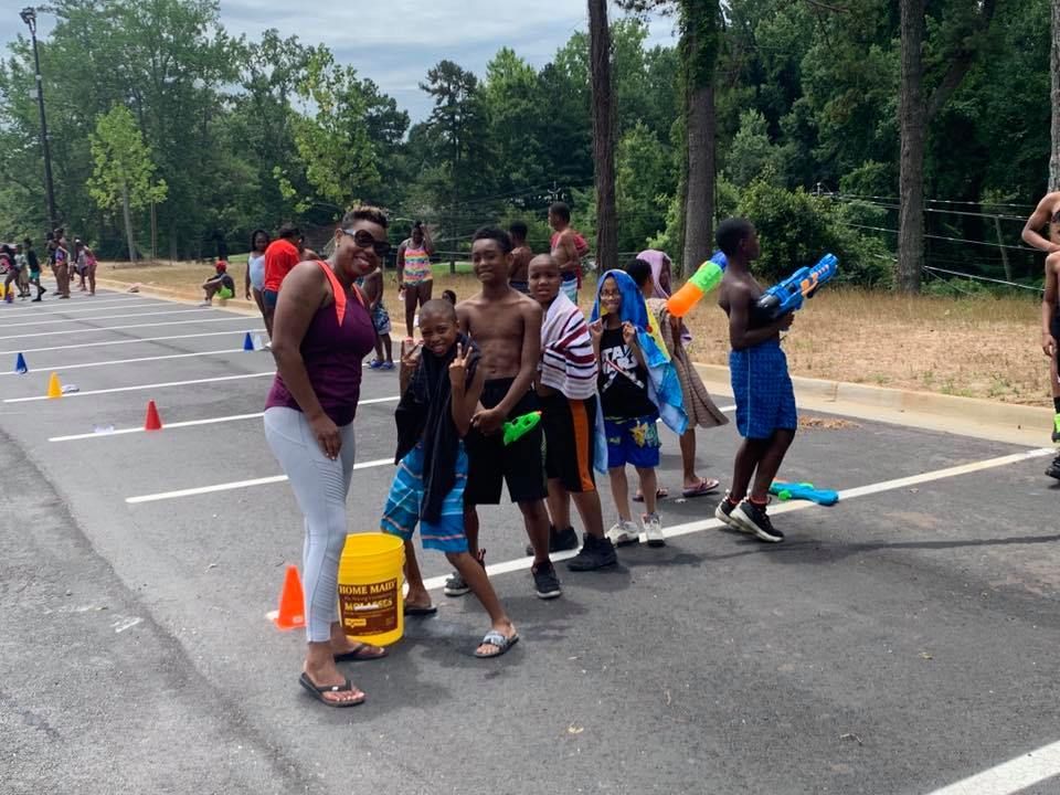 A group of children and an adult standing in a parking lot with water toys, bright orange traffic cones, and trees.