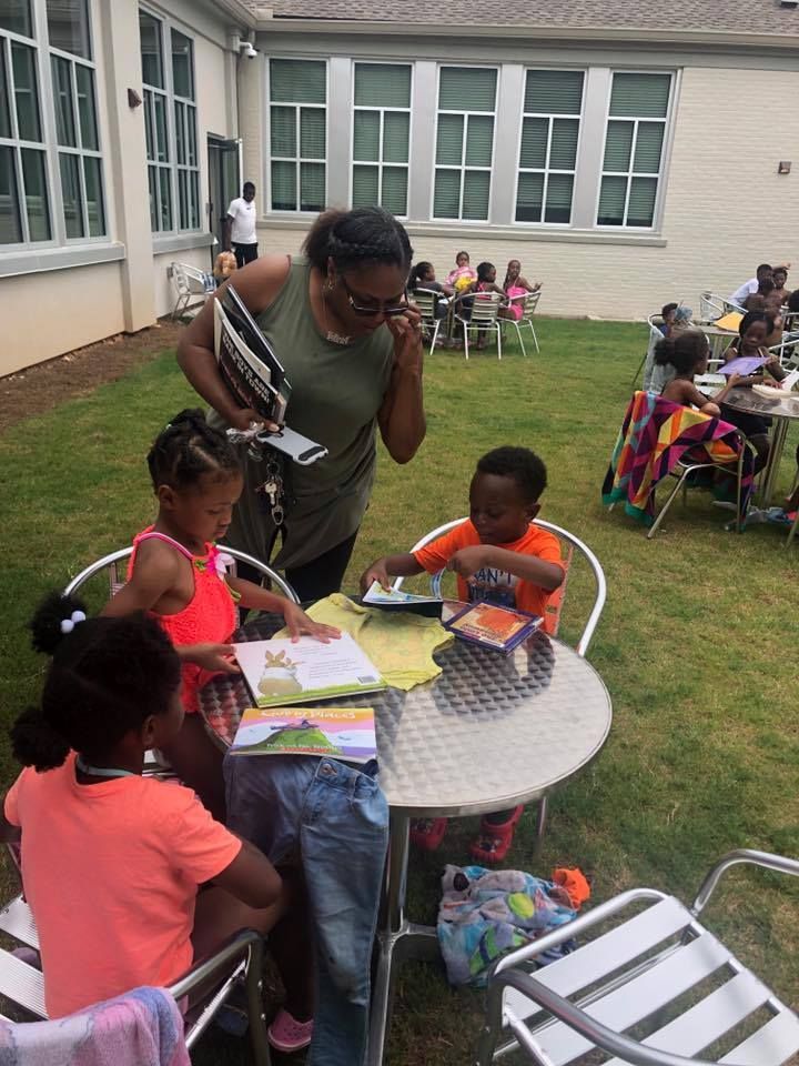 A woman stands over a group of children reading books at a metal outdoor table on a grassy patio.